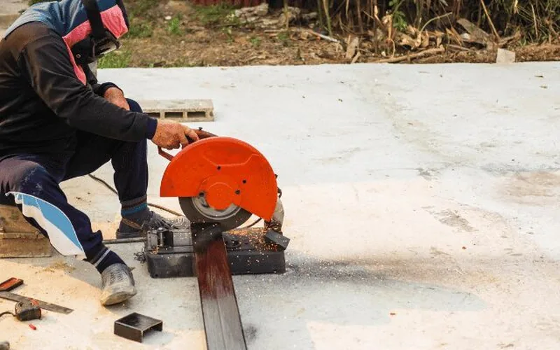 worker using an abrasive metal-cutting saw to cut a steel bar, sparks flying from the cut
