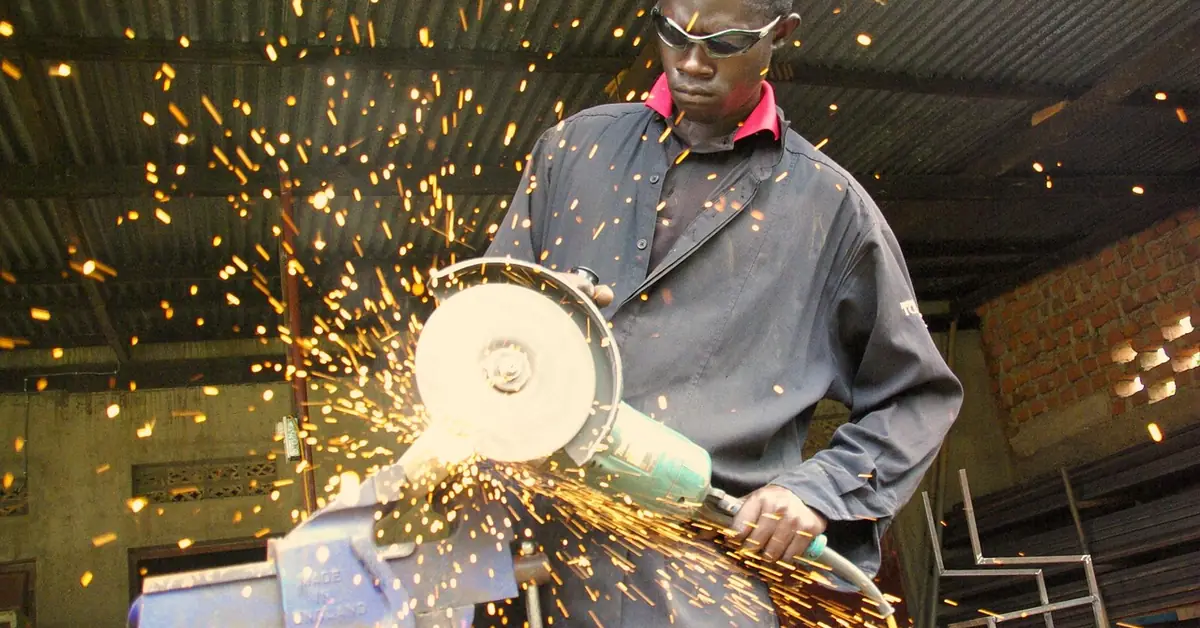 worker wearing safety glasses cutting a steel bar clamped in a vise, sparks flying from the blade