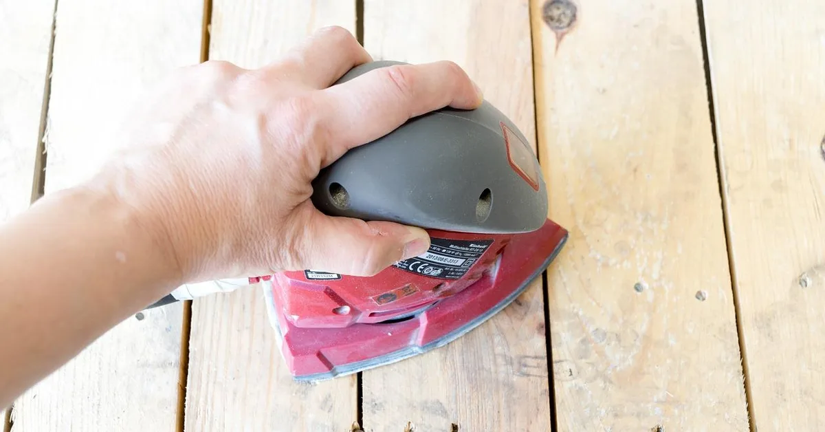 Hand holding a detail sander over bare plank boards to smooth a repaired area