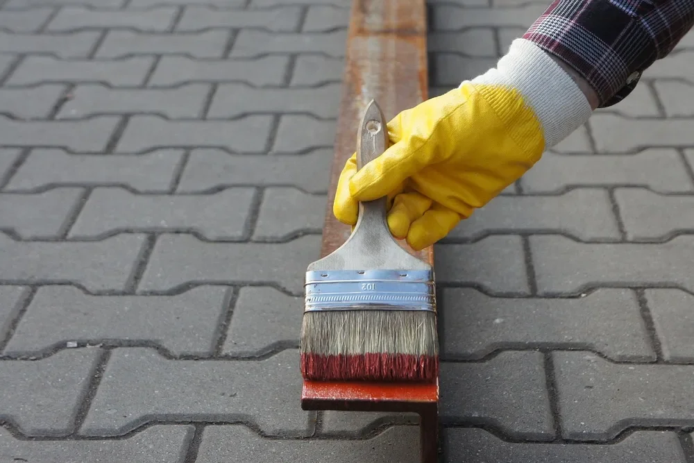 Gloved hand holding a paintbrush over a metal bar, showing brush application of a coating