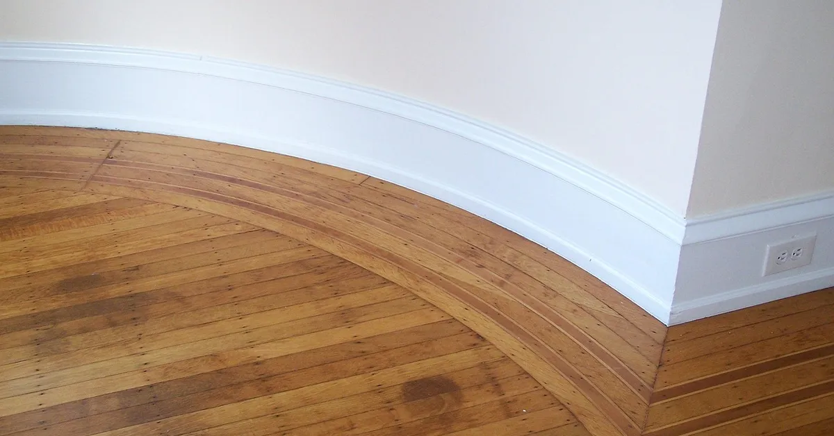 White baseboard meeting a hardwood floor at an inside corner, where quarter round is nailed to the baseboard