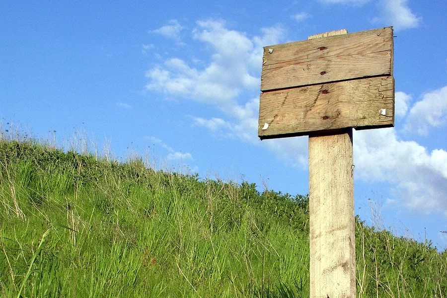 Plain wooden sign board nailed to a post standing in tall grass under a blue sky