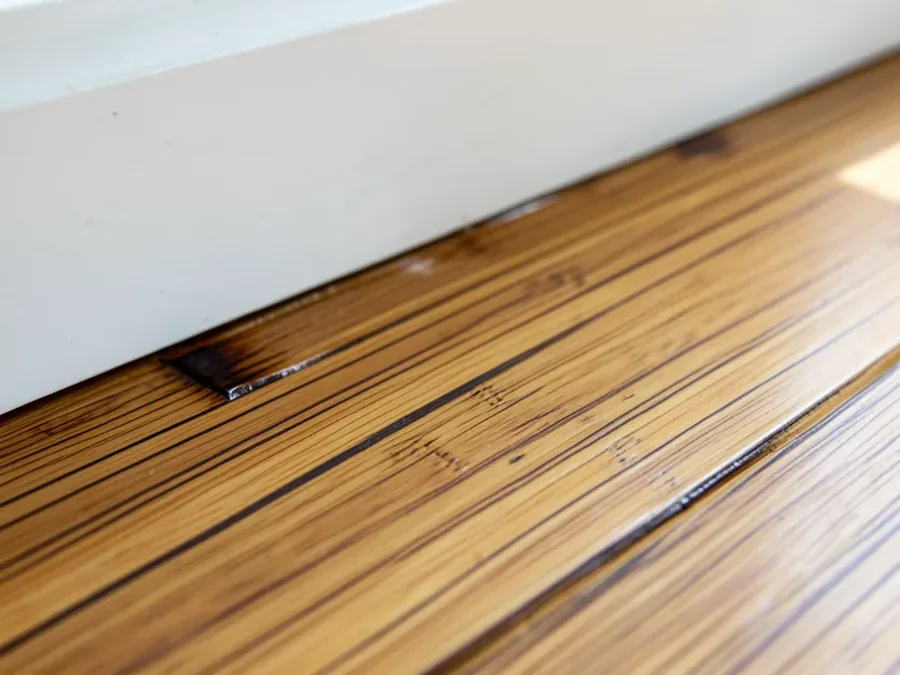Close-up of a strand-woven bamboo floor plank near a baseboard showing slight cupping and discoloration from moisture damage