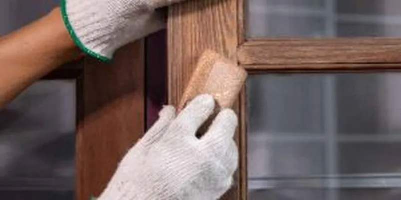 Gloved hands sanding a wood window frame edge with a sanding block