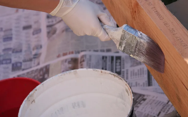 Gloved hand brushing paint onto a wood board next to an open paint bucket