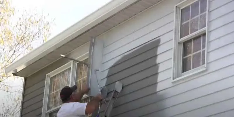 Man spray-painting cedar siding with a sprayer while standing on a ladder