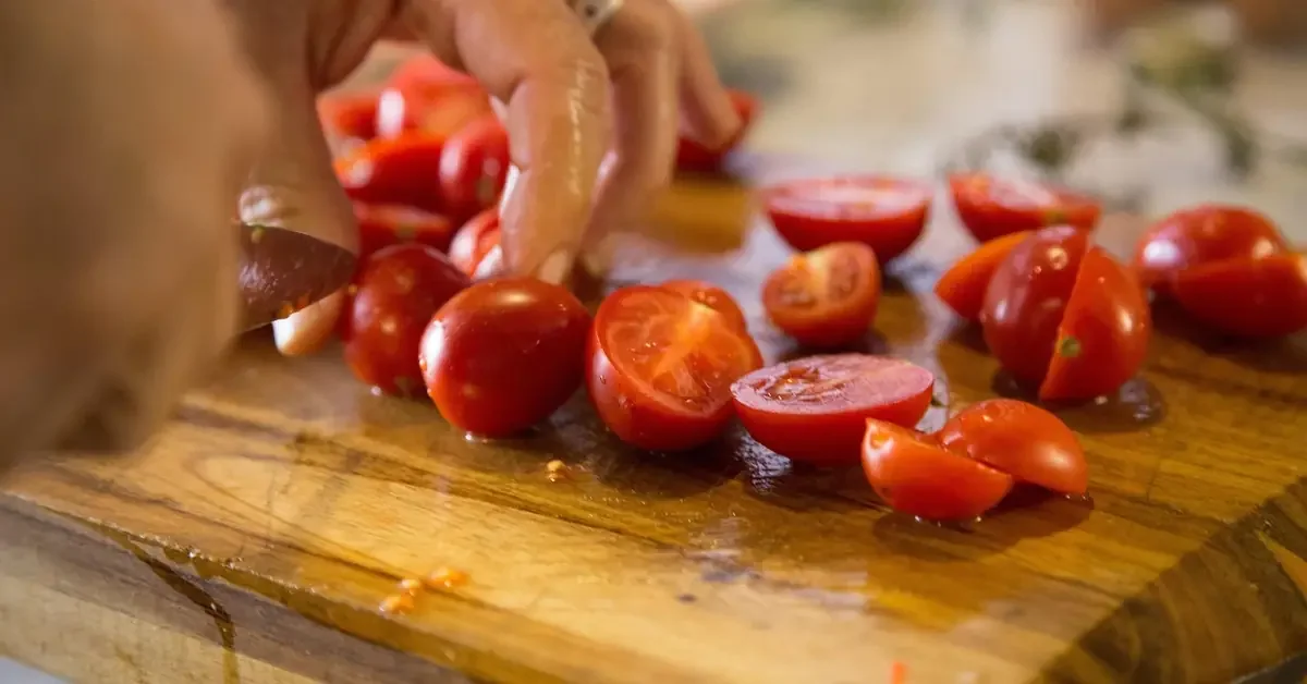Hands slicing cherry tomatoes on a wooden cutting board with warm grain