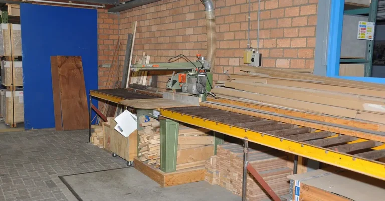 A green radial arm saw with an overhead blade arm set up in a woodworking shop, cutting lumber to length