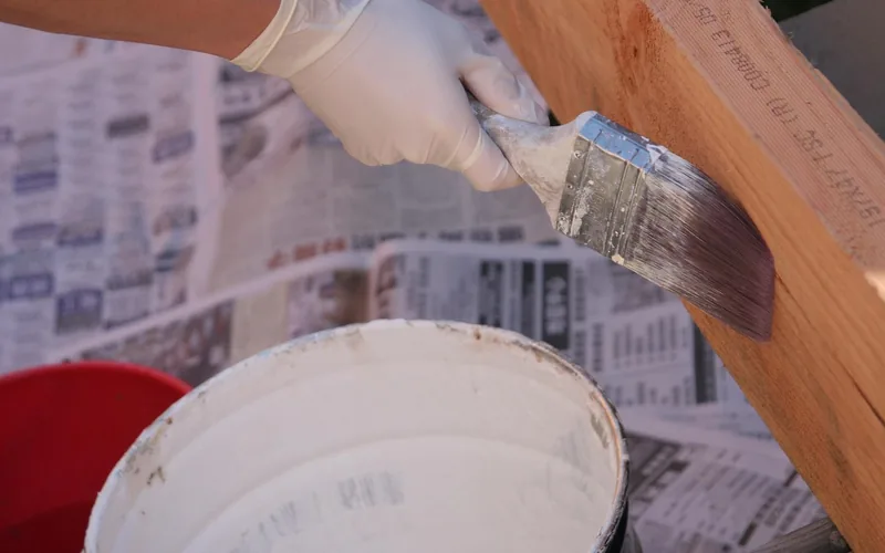 Gloved hand brushing white paint onto a wood board over a drop cloth