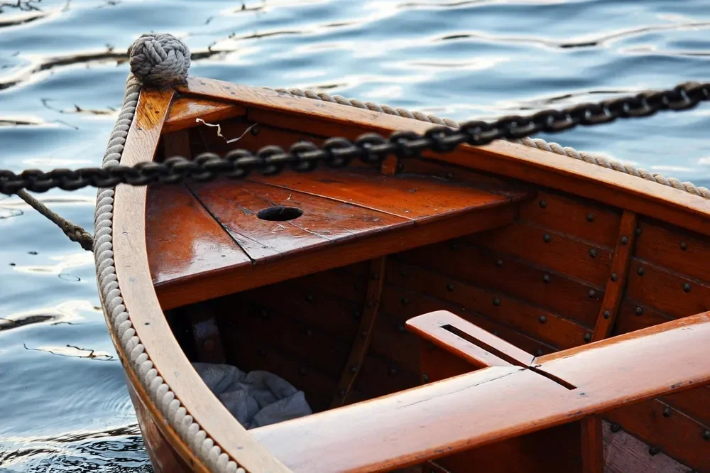 Bow of a varnished wooden rowboat with planked sides and a rope rail on the water