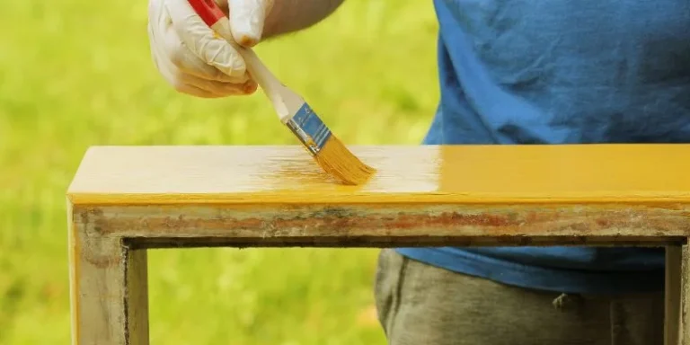 Applying clear lacquer finish with a brush to a wood surface, showing the wet amber tone before it cures