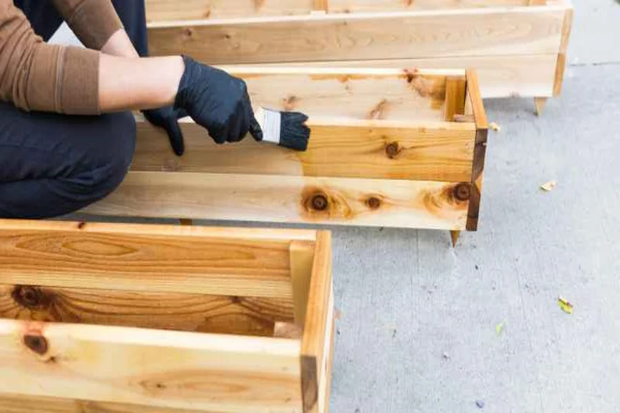 Person in gloves applying stain to a cedar planter box, wood grain absorbing the stain evenly