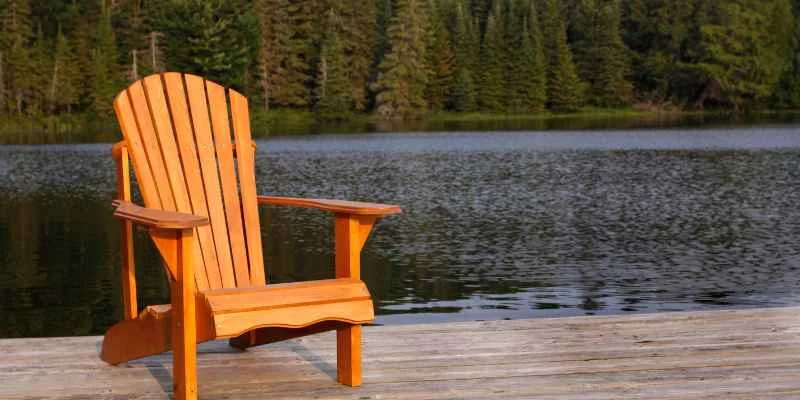 single wood Adirondack chair on a dock facing a forested lake