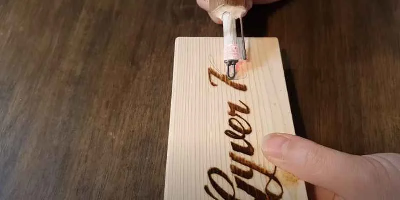 Hand-held wood burning pen tracing cursive lettering into a pine board