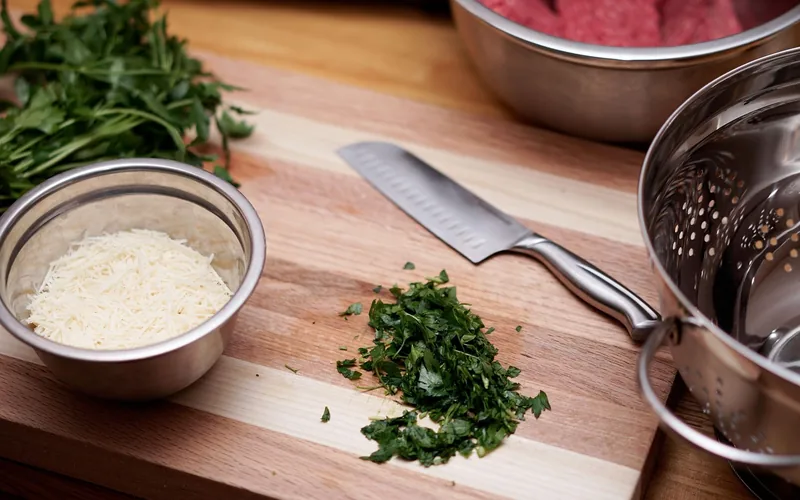 chef knife resting on a wood cutting board next to shredded parmesan and chopped herbs