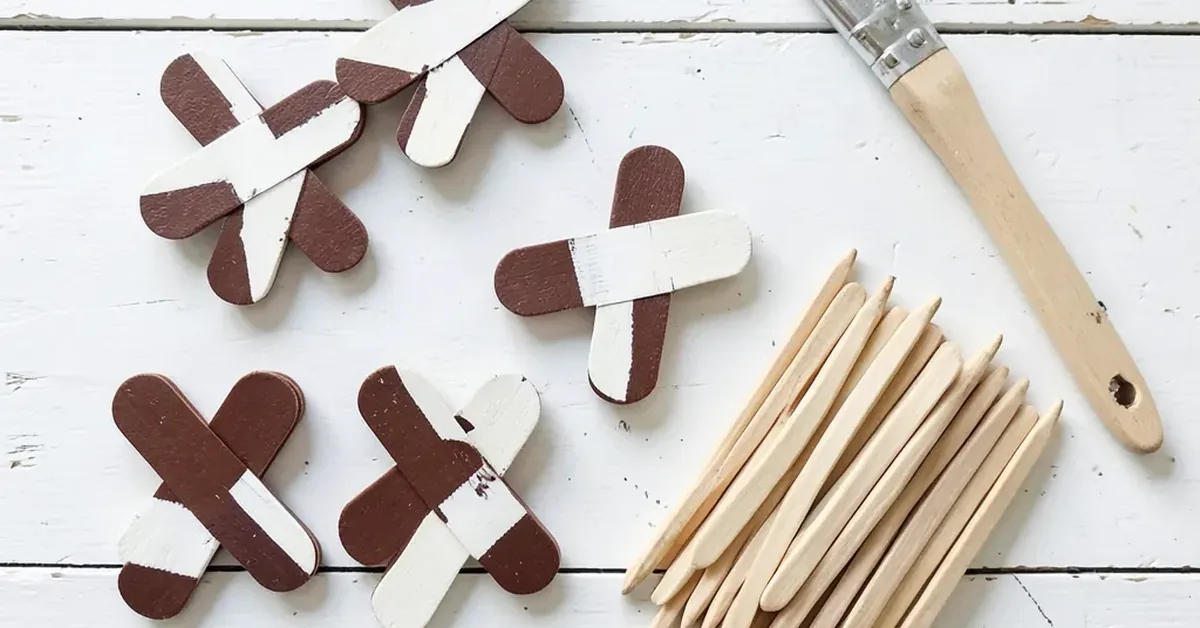 Five finished popsicle stick cross ornaments painted brown and white next to unpainted craft sticks and a paintbrush