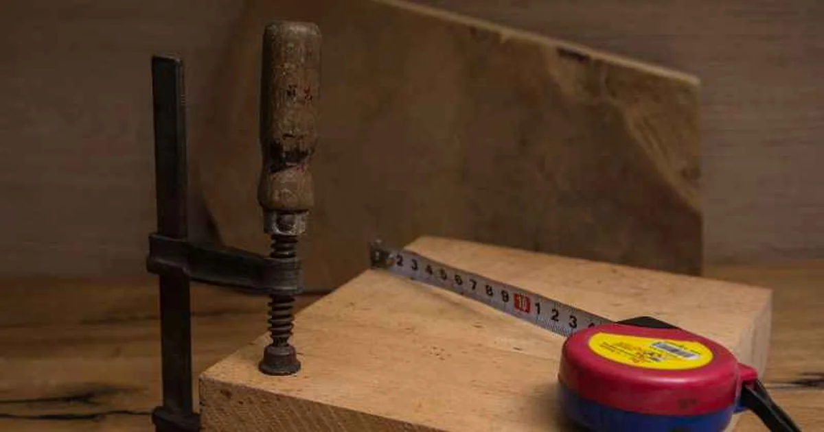 Bar clamp holding a board with a tape measure laid across it, used to plan how much extra reach a clamp extension needs
