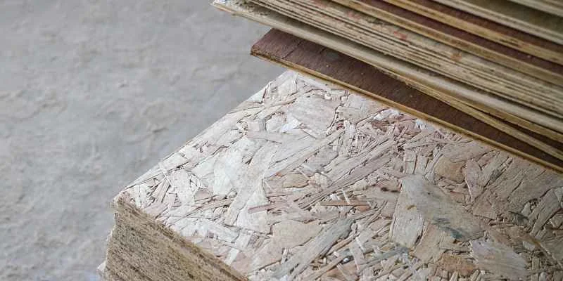 Close-up edge view of an OSB panel next to stacked plywood sheets showing the strand versus veneer layer structure