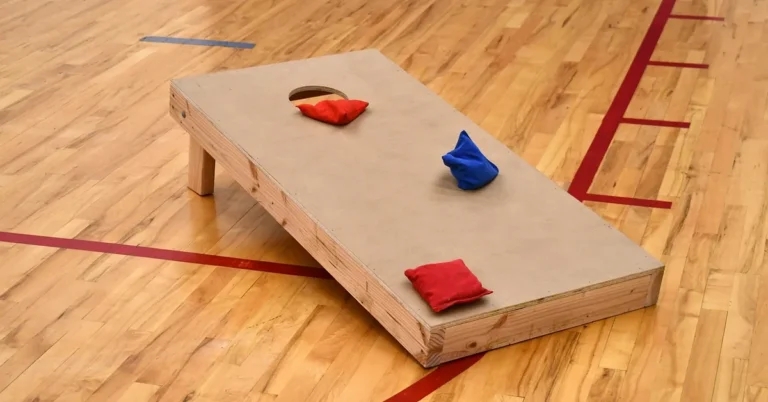 Wood frame bean bag toss board with a 6 inch hole and two bags on a gym floor
