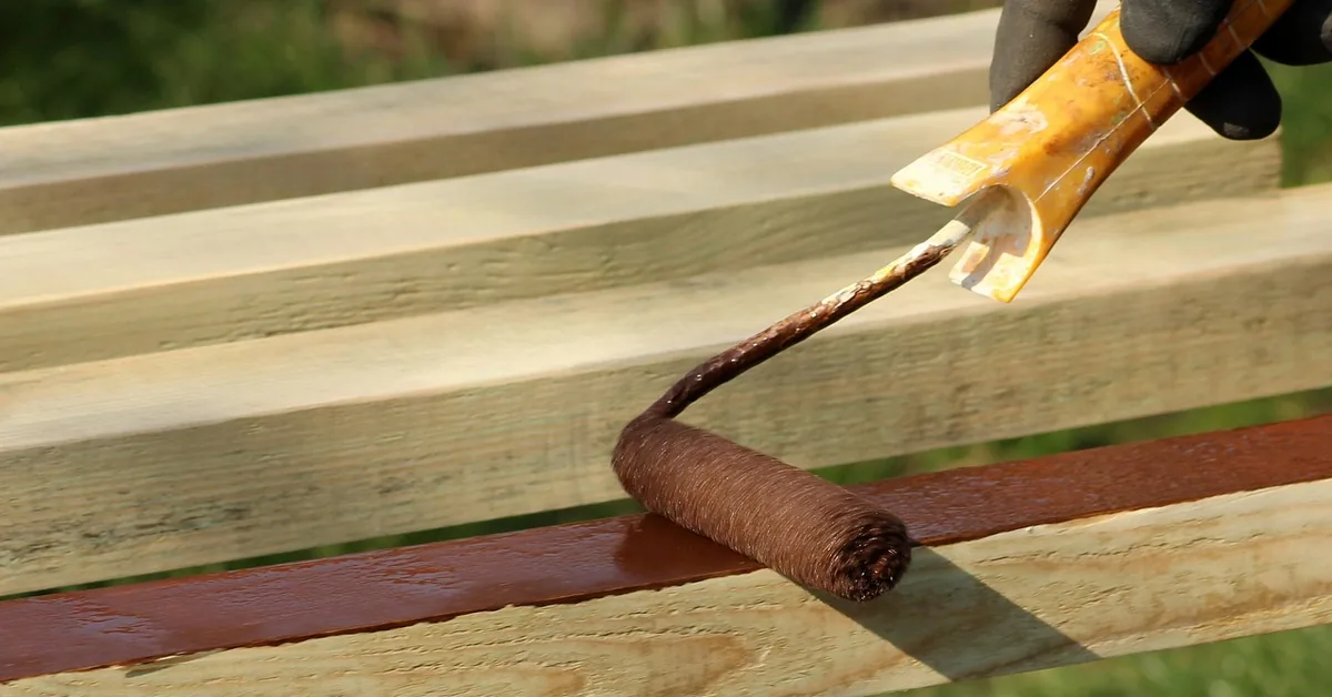 Gloved hand using a paint roller to apply flat finish paint to a pressure-treated wood fence rail outdoors