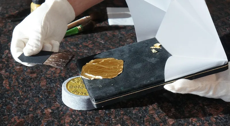 Gloved hands lifting a sheet of gold leaf with a gilding brush