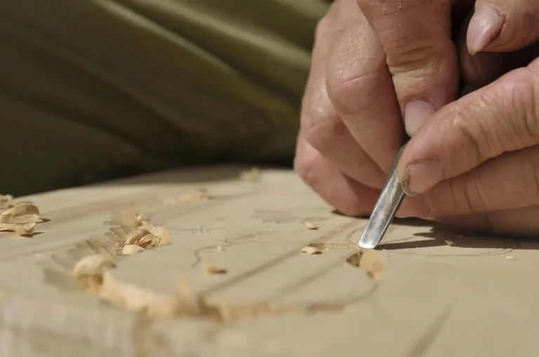 Hand pushing a carving gouge through pale wood, leaving curled shavings