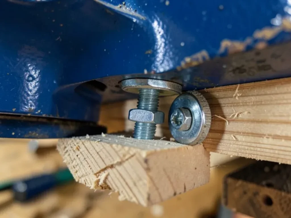 Through-bolt, washer, and nut being tightened from underneath a wooden workbench to secure a woodworking vise