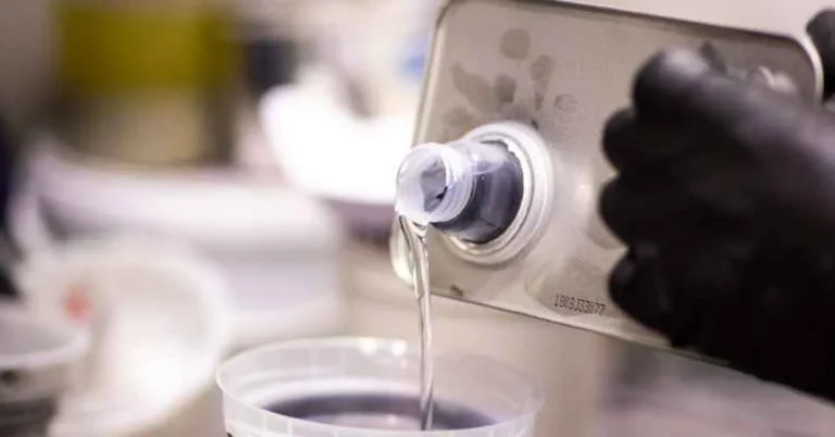 Gloved hand pouring clear paint solvent from a metal can into a measuring cup