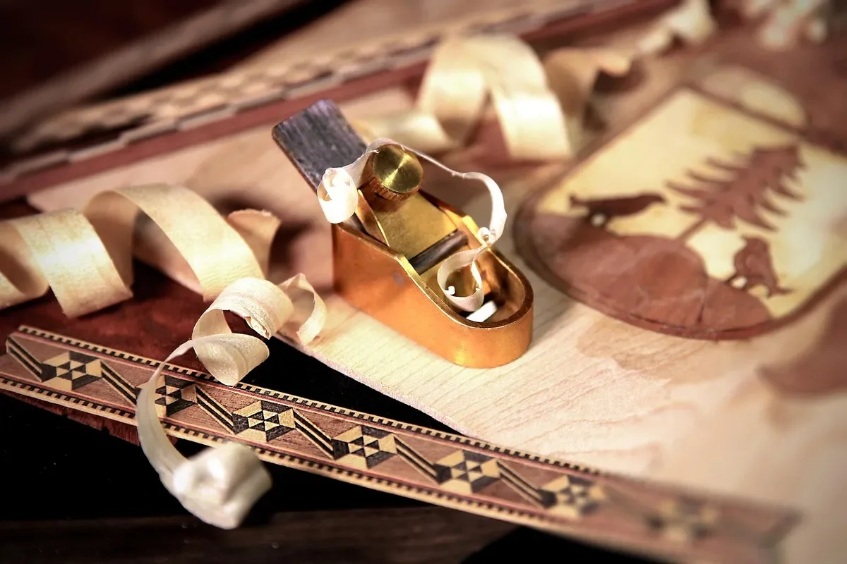 Small brass hand plane and pale shavings on thin wood veneer with marquetry inlay