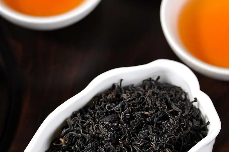 Dry black tea leaves in a white dish beside cups of brewed black tea