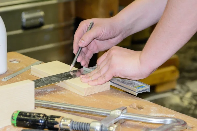 Hand marking a cut line on a wood block with a pencil and metal square, a bar clamp and glue bottle on the bench
