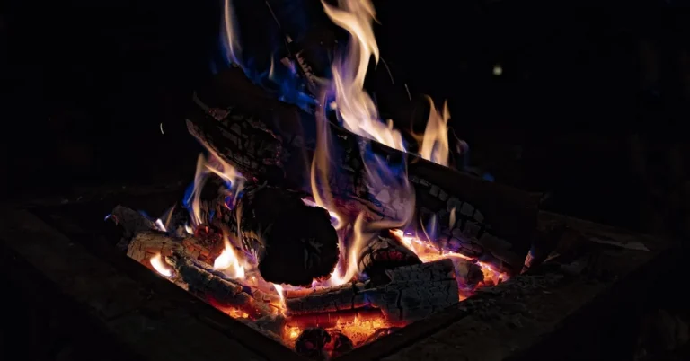 Stacked firewood burning inside a metal fire pit at night with tall orange flames