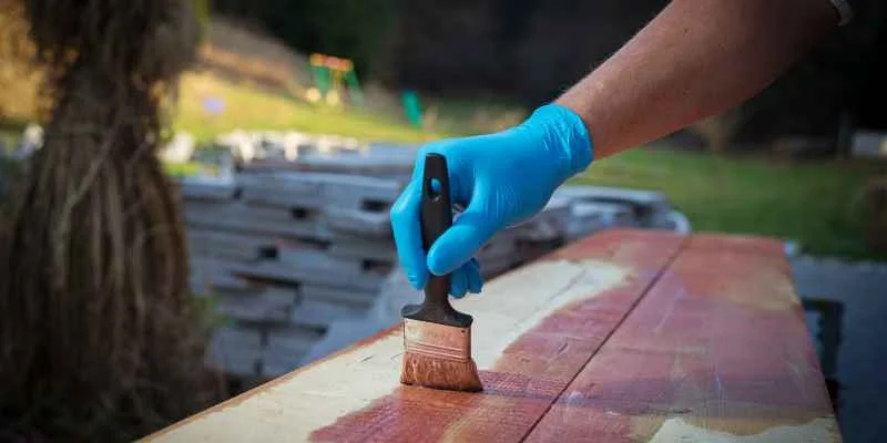 gloved hand applying wood stain to an outdoor wood plank