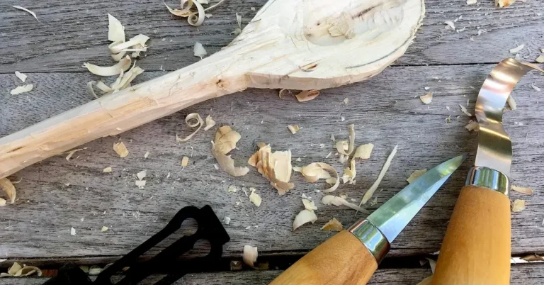 Two carving knives beside a half-carved wooden spoon and wood shavings on a workbench