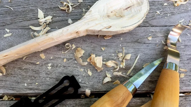 Whittling knives and wood shavings on a workbench beside a carved wooden spoon