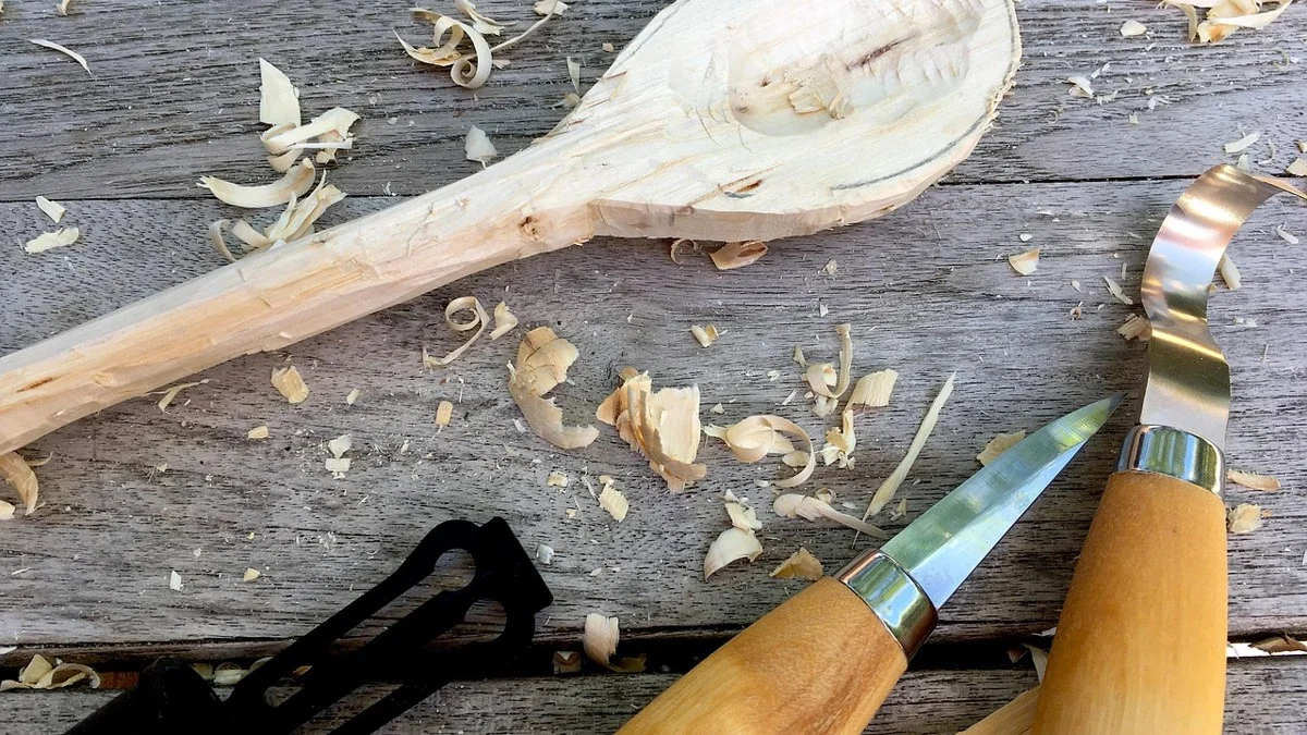 Whittling knives and wood shavings on a workbench beside a carved wooden spoon