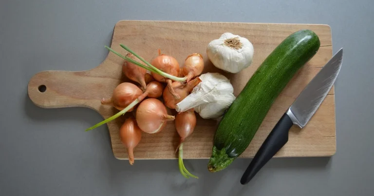 raw shallots, garlic bulbs, and a zucchini on a wooden cutting board with a chef knife