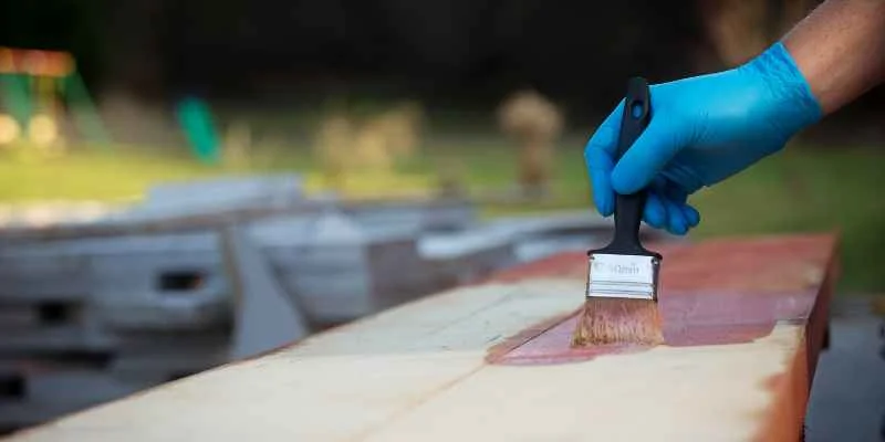 person wearing nitrile gloves applying dark wood stain to a board outdoors with a paintbrush