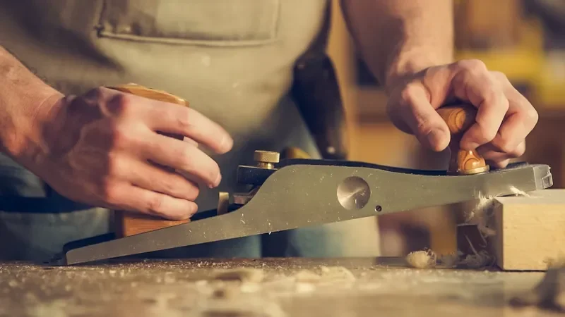 Hands pushing a hand plane along a board on a workbench with shavings