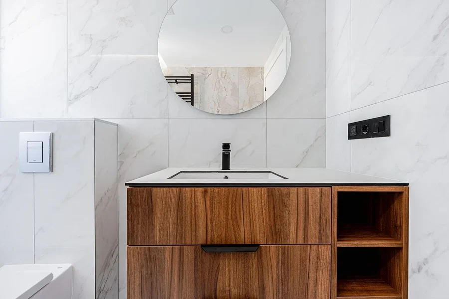 Round mirror mounted on a tiled wall above a walnut-look wood vanity