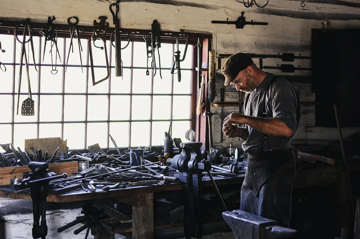Craftsman in a long work apron standing at his workbench in a tool-filled shop