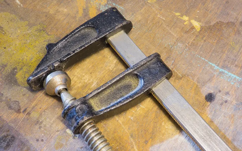 Steel bar clamp lying on a worn wooden workbench, the kind used to hold a warped board flat while it dries