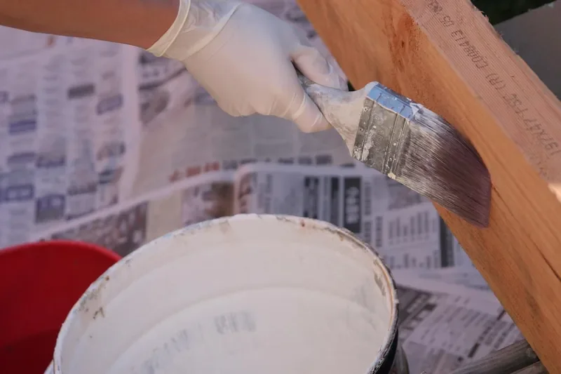 Gloved hand brushing white primer onto a wood board from a paint bucket Gloved hand brushing white primer onto a wood board from a paint bucket