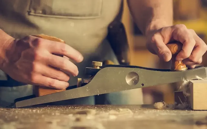 Hands using a hand plane to shave a thin layer off a wood board Hands using a hand plane to shave a thin layer off a wood board