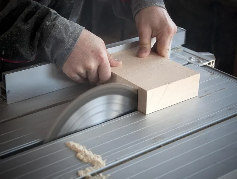 Close-up of a table saw blade cutting a block of wood, hands guiding the workpiece