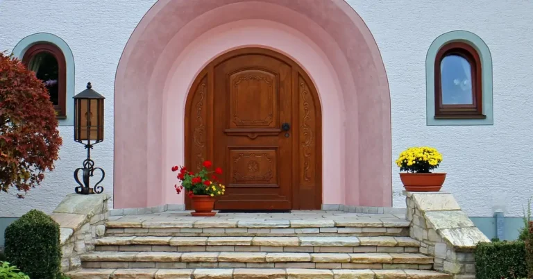 Arched natural wood teak-style main door with stone entryway and potted flowers