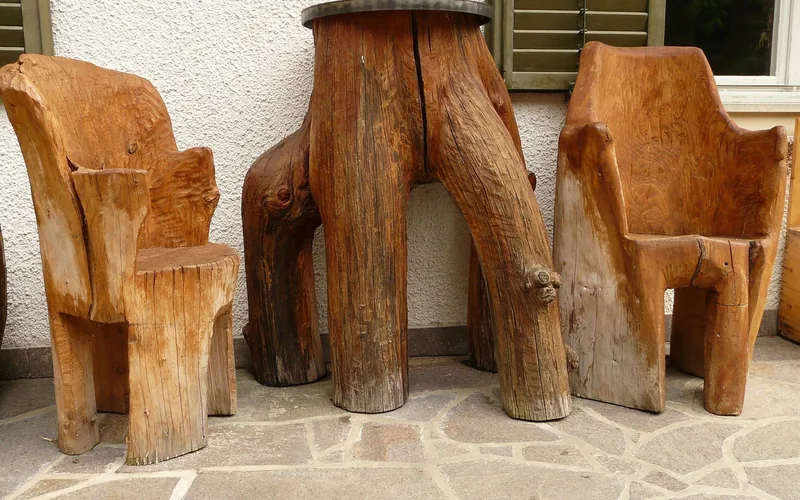 Unfinished wood chairs and a table standing on a stone patio next to a wall