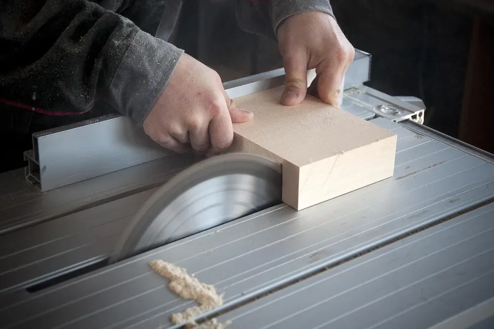 Hands feeding a wood block past a table saw blade with sawdust on the table