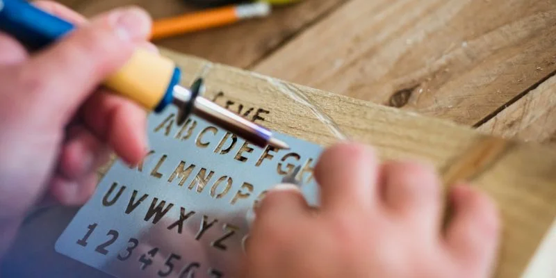 Tracing a wood burning pen tip through a metal alphabet stencil to burn straight even letters