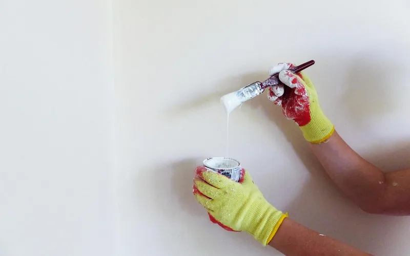 gloved hands dipping a paintbrush into a can of white paint near a prepped wall gloved hands dipping a paintbrush into a can of white paint near a prepped wall